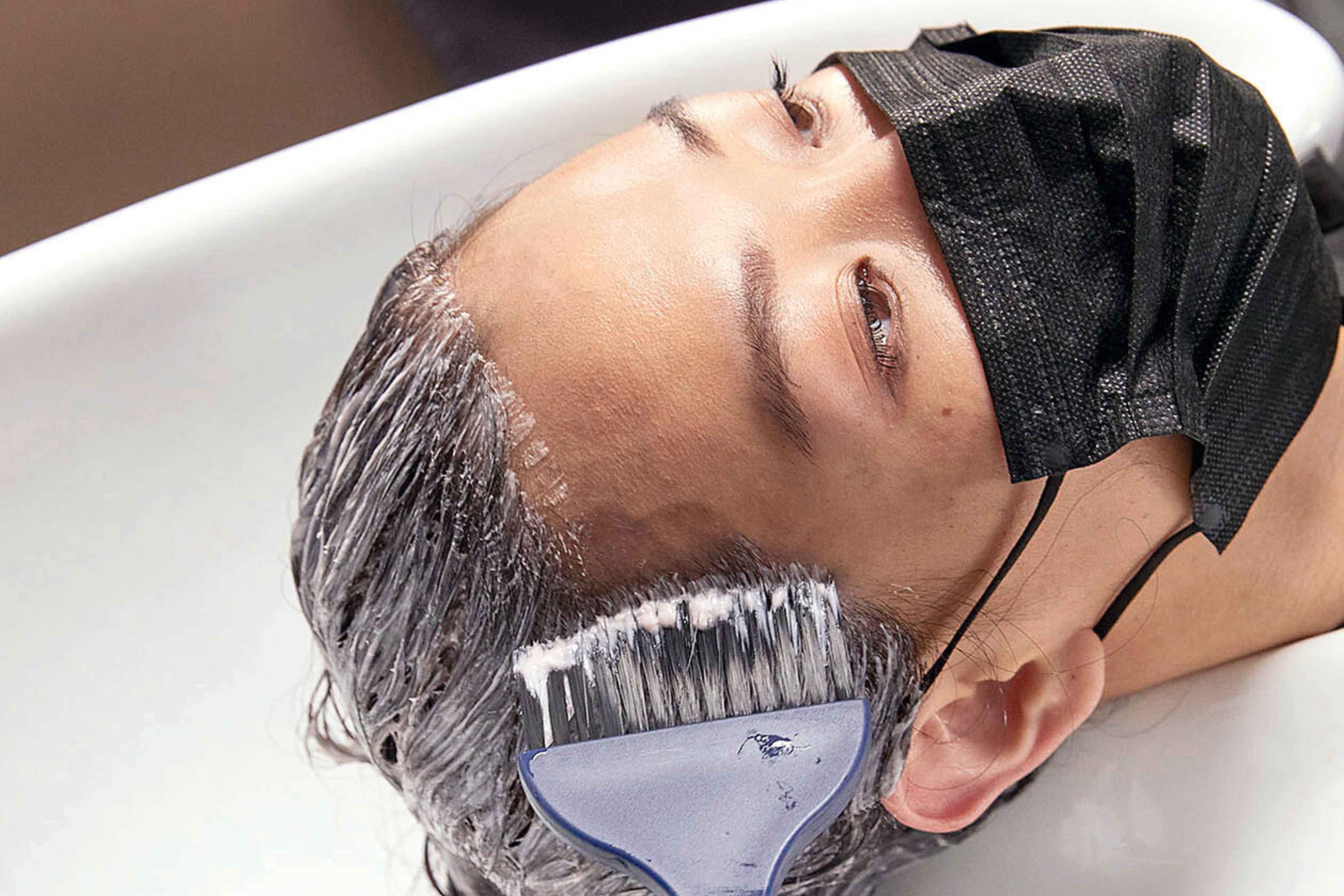 Einer Frau mit Atemschutzmaske werden an einem Waschbecken mit einem blauen Pinsel die Haare gefärbt.