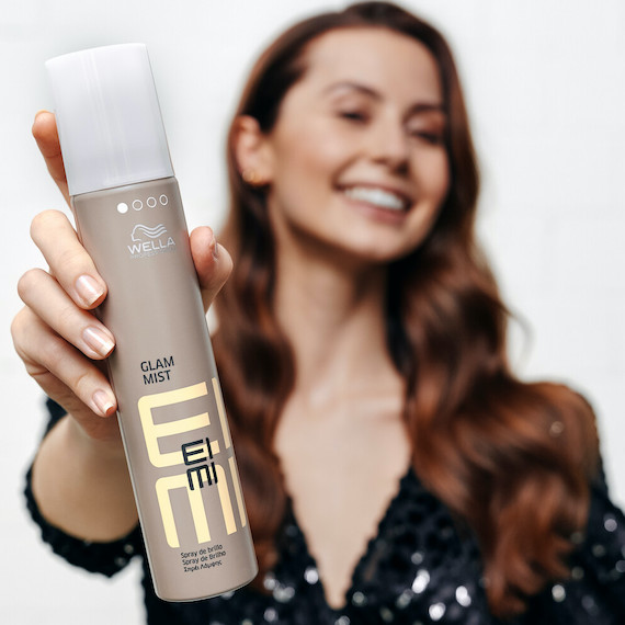 Ein Model mit langem, gewelltem braunem Haar hält eine Flasche EIMI Glam Mist von Wella Professionals in der Hand