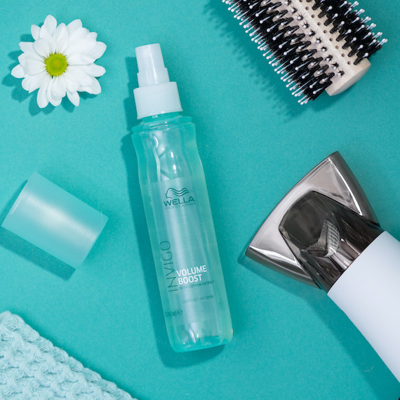 Eine blaue Flasche Wella Professionals Invigo Volume Boost Hair Mist umgeben von einer Margeritenblüte, einer Haarbürste und einem Föhn
