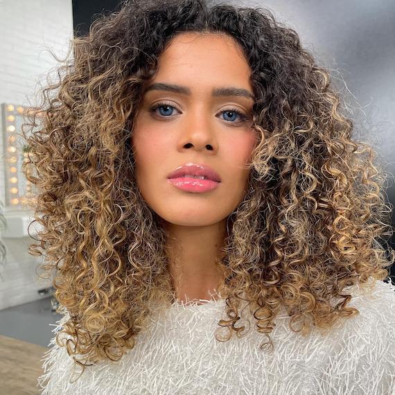 Eine Frau mit schulterlangen Locken und blonder Balayage, kreiert mit Wella Professionals, blickt in die Kamera.