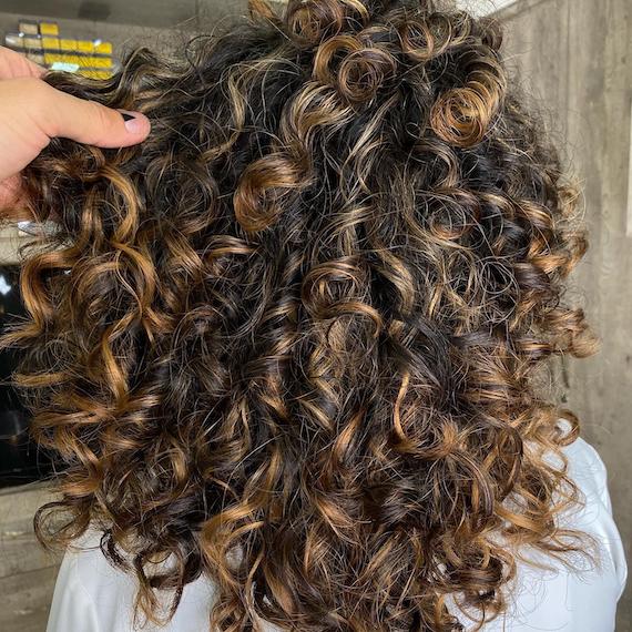 Hinterkopf einer Frau mit dunkelbraunem, lockigem Haar und chunky, karamellblonder Balayage, kreiert mit Wella Professionals.