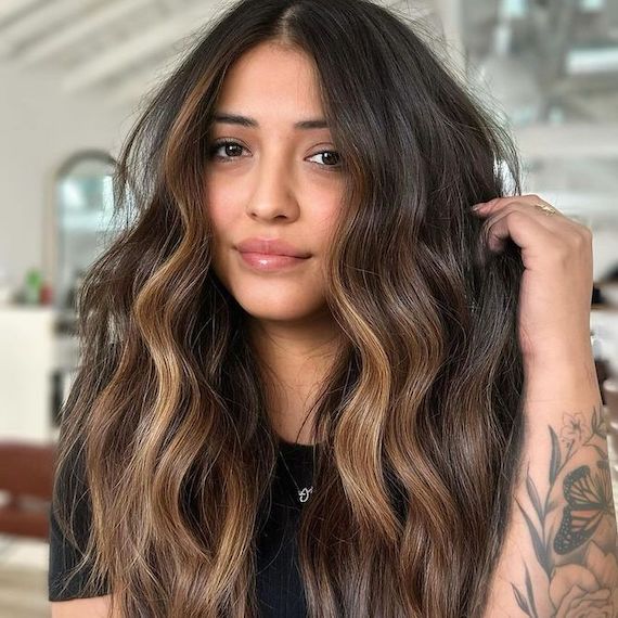 Model with long, wavy, brunette hair and a golden blonde balayage faces camera.