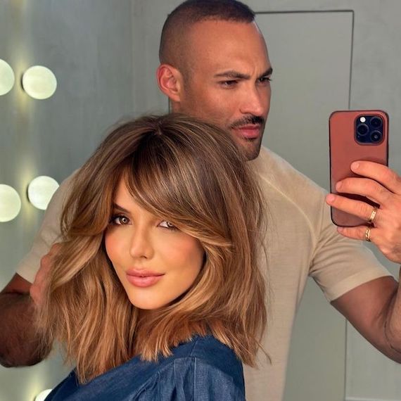 Model with shoulder-length, brown hair and blonde highlights, featuring grown-out curtain bangs.