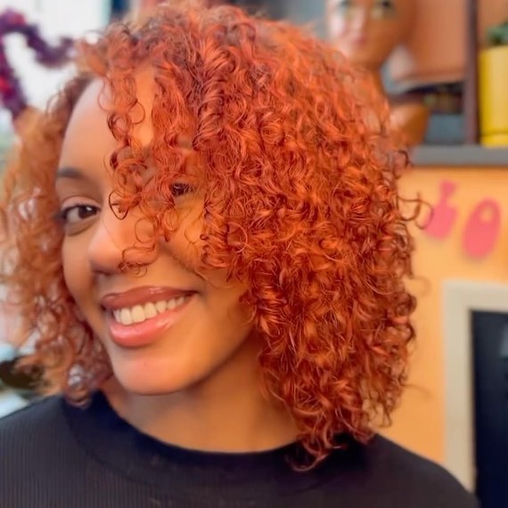 Model with shoulder-length, copper red, curly hair and long side bangs.