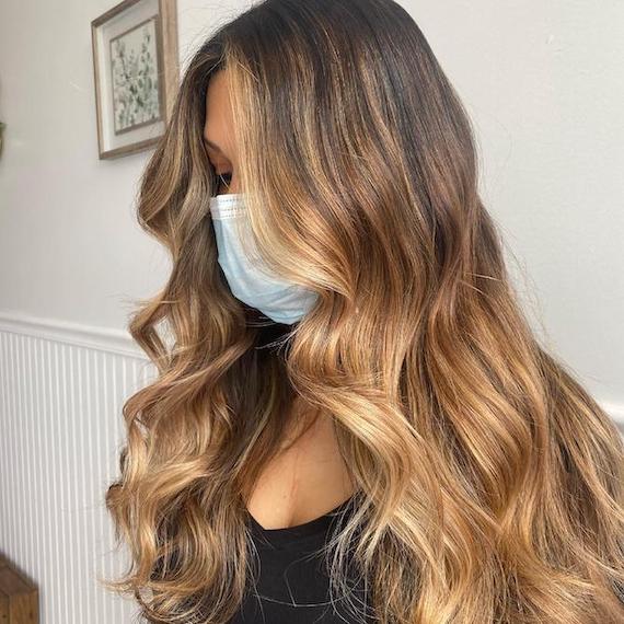 Model with long brown hair and blonde balayage wears a face mask and looks down