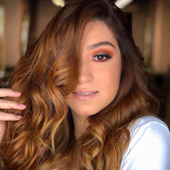 Model faces the camera with half of their face concealed by long, caramel brown hair. Their hand holds up a section of hair.