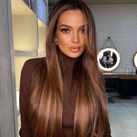 Model with long, dark brown, highlighted hair stands in front of a mirrored wall. They look straight at the camera.