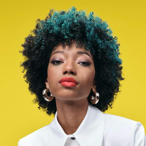 Model with black coily hair and teal highlights looks directly into the camera.