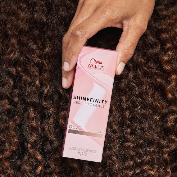 Model with glossy, brown, curly hair holds a box of SHINEFINITY.