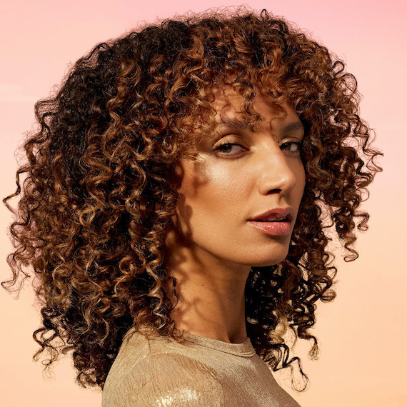 Model with shoulder-length, curly brown hair and sun-kissed bronze highlights.