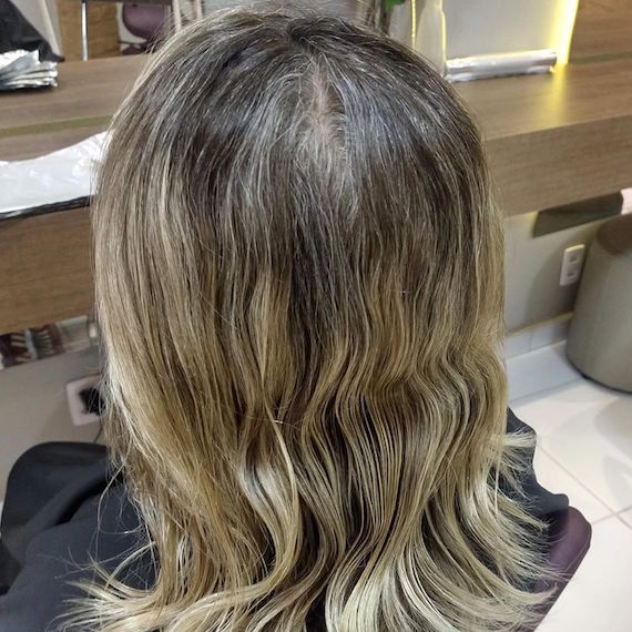 Hinterkopf einer Frau mit gewelltem, blondem Haar und grauen Ansätzen.
