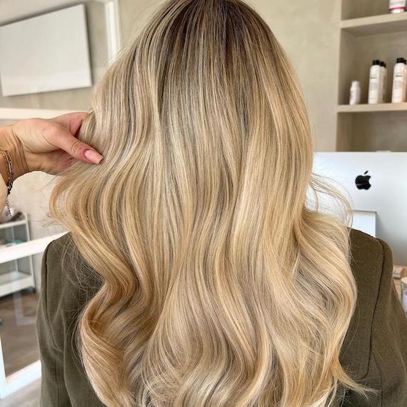 A person with long, honey blonde babylights sits in a salon with her back to the camera to show off her newly coloured hair