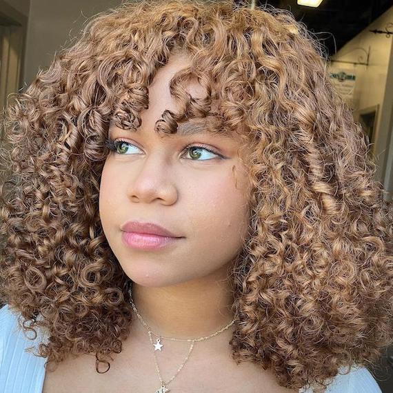 Headshot of a person with deep honey blonde chunky highlights on curly hair and hazel green eyes