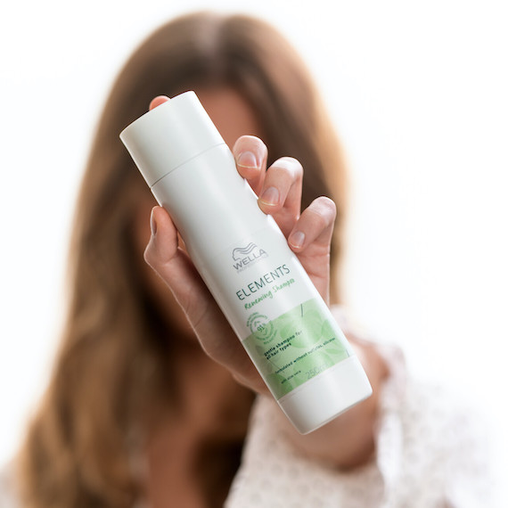 Woman holds Wella Professionals Elements Renewing Shampoo in front of her face.