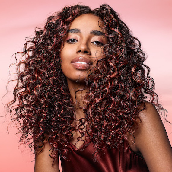 Model with long, dark brown, curly hair featuring coral highlights.
