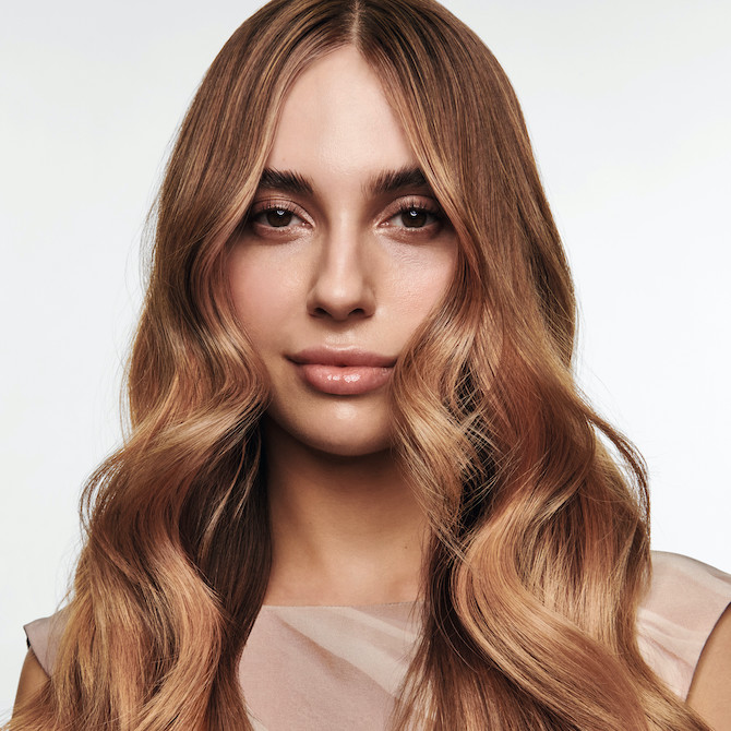 Model with long, loosely curled, light brown hair featuring rose gold balayage.