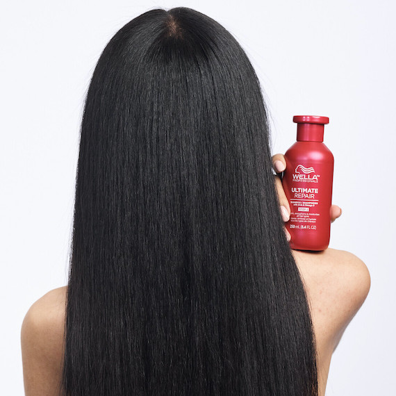Model with long, straight, dark brown hair holds a bottle of ULTIMATE REPAIR Shampoo.