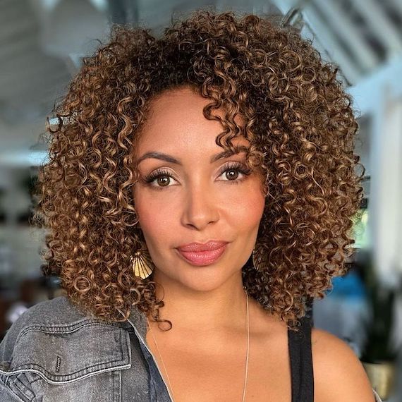 Model with glossy, brown, curly hair and caramel highlights looks directly into the camera.