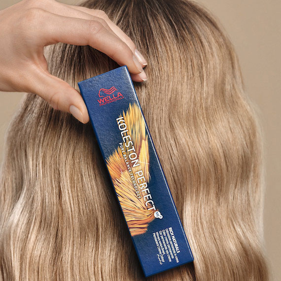 Hand holds a box of Koleston Perfect blonde hair color by Wella Professionals against the back of a head