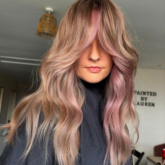 Model with long, wavy, ash blonde hair, grown-out bangs and purple-pink face-framing highlights.