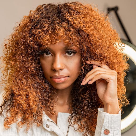 Model with dark brown curly hair featuring a copper peach shade through the lengths.