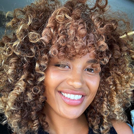 Model with curly, brown hair featuring honey blonde highlights and a touch of peach around the face frame.