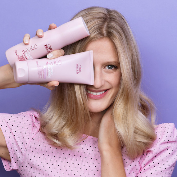 Model with long blonde hair holds a bottle of Wella INVIGO Blonde Recharge shampoo and conditioner