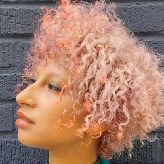 Model mit kurzem, lockigem, pastellrosa und pfirsichfarbenem Haar schaut zur Seite.