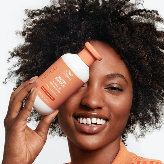 Model with black, curly hair holds up a bottle of INVIGO Nutri-Enrich Deep Nourishing Shampoo.