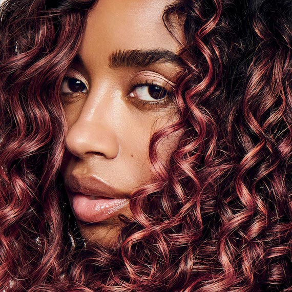 Model with dark brown, curly hair featuring glossy coral highlights.