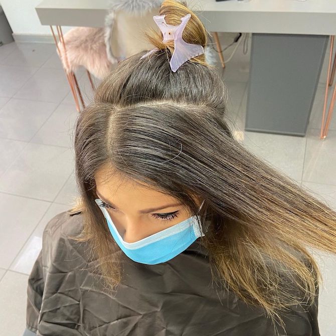Woman with long dark hair and grey roots, sitting in a salon chair.