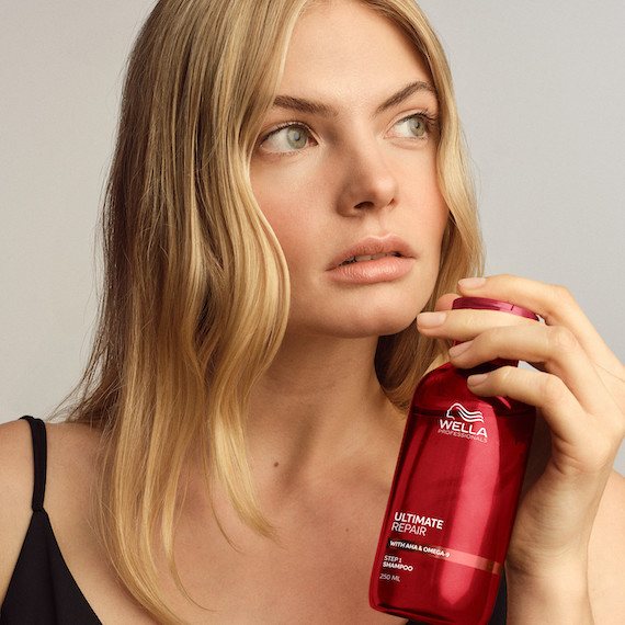 Model with honey blonde, straight hair holds up a bottle of ULTIMATE REPAIR Shampoo.